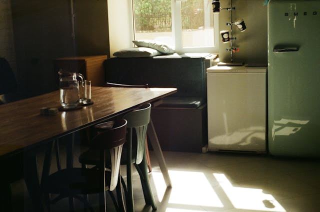 A sunlit room with a wooden table, chairs, and a refrigerator.